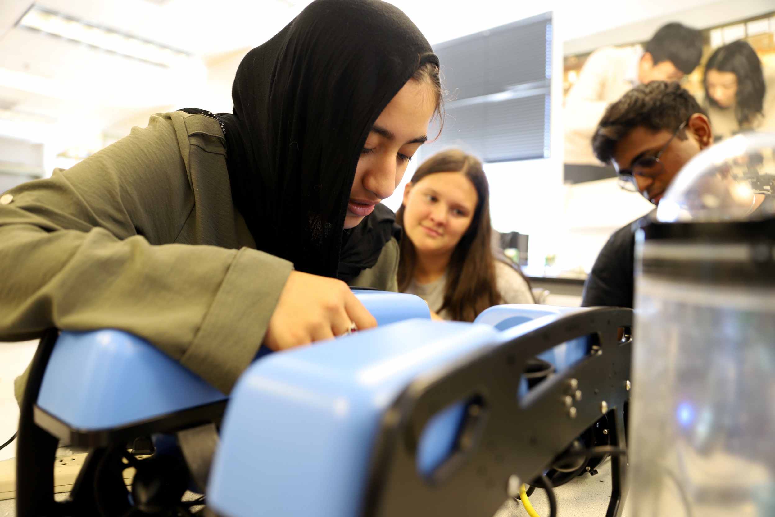 A student leans in closely to examine or adjust a small blue robotic device, while two classmates look on in a university lab setting.