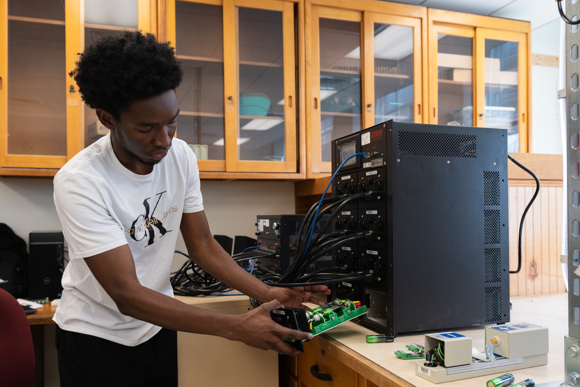 A student works with a circuit board and large electronic equipment rack in a university engineering or computer science lab.