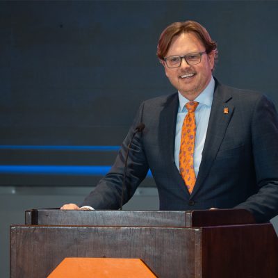 Jeff Stoecker stands at a wood podium, smiling, wearing a dark suit and orange patterned tie with a Syracuse University lapel pin.