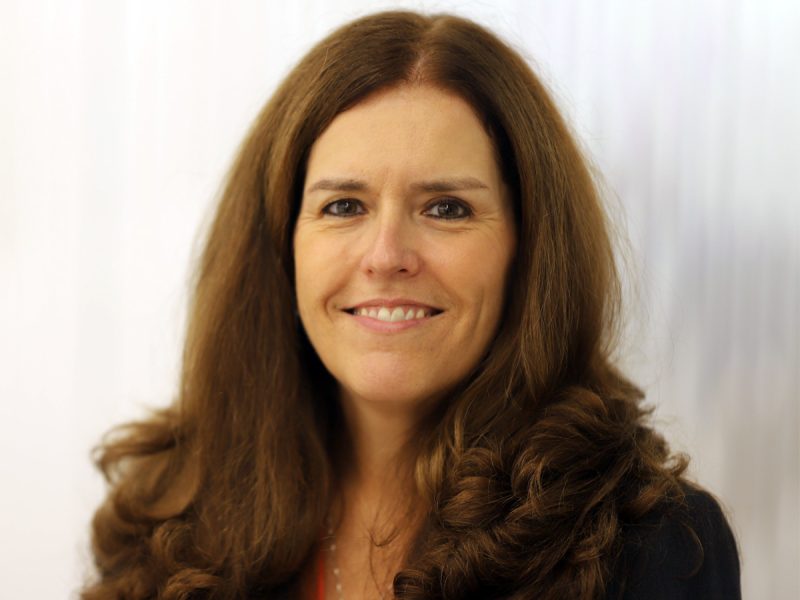 A person smiles for a headshot wearing a dark blazer and orange blouse.