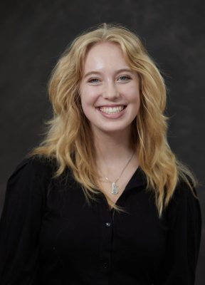 Professional headshot of a smiling woman with wavy blonde hair, wearing a black button-up shirt and a silver pendant necklace, against a dark gray background.
