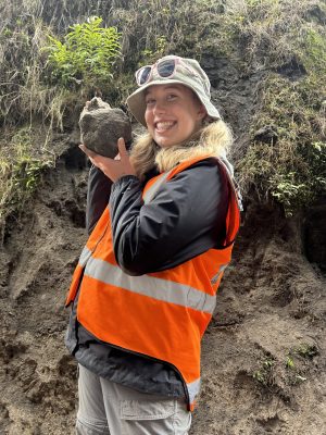 Person in an orange safety vest and bucket hat smiling while holding up a chunk of volcanic rock, standing in front of a volcanic soil embankment with vegetation.
