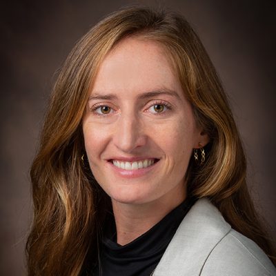 Headshot of a smiling woman with long reddish-brown hair wearing a light gray blazer and black top against a dark brown background.