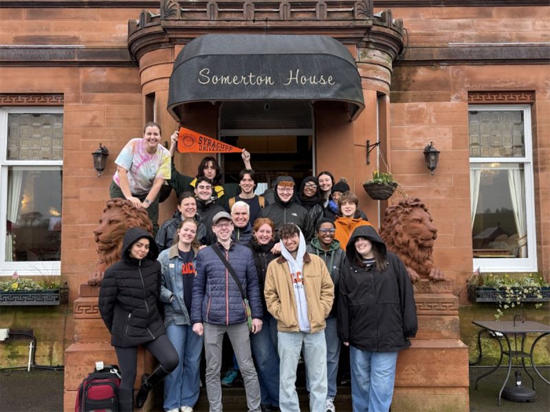 A group of Syracuse University students and faculty pose outside Somerton House, a red sandstone building with stone lion sculptures, one person holding an orange SU pennant.
