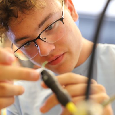 A student concentrates closely while using a small tool to work on wiring or an electronics component in a lab setting.