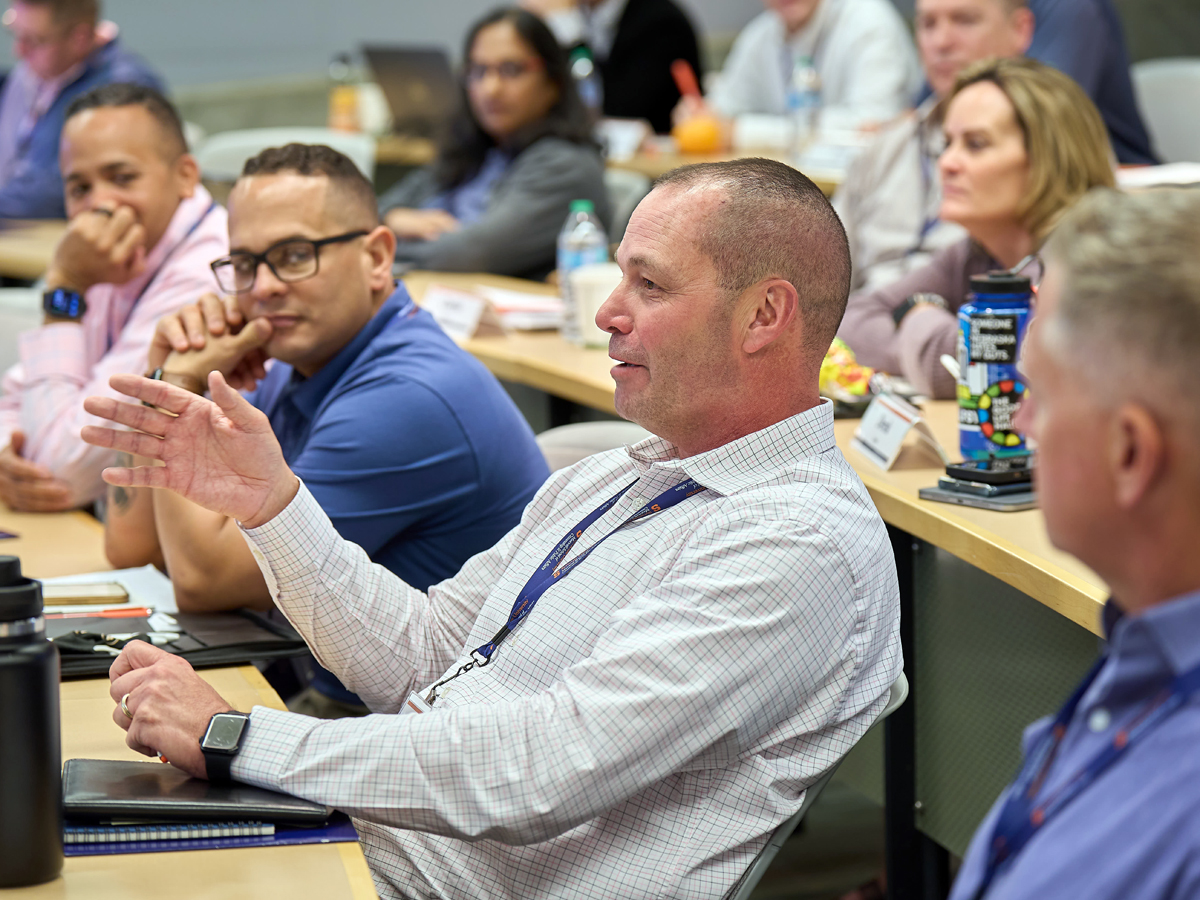 A man gestures while speaking during a classroom discussion, surrounded by engaged participants.