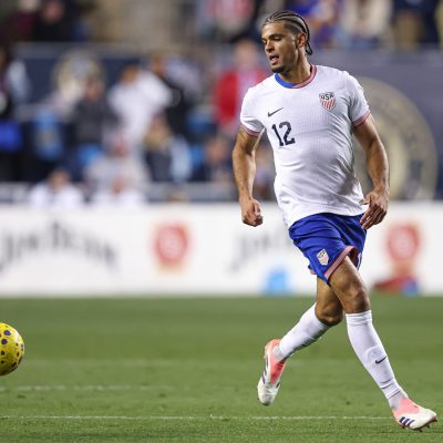A person dribbles the ball in a USMNT match wearing the team's white and blue uniform.
