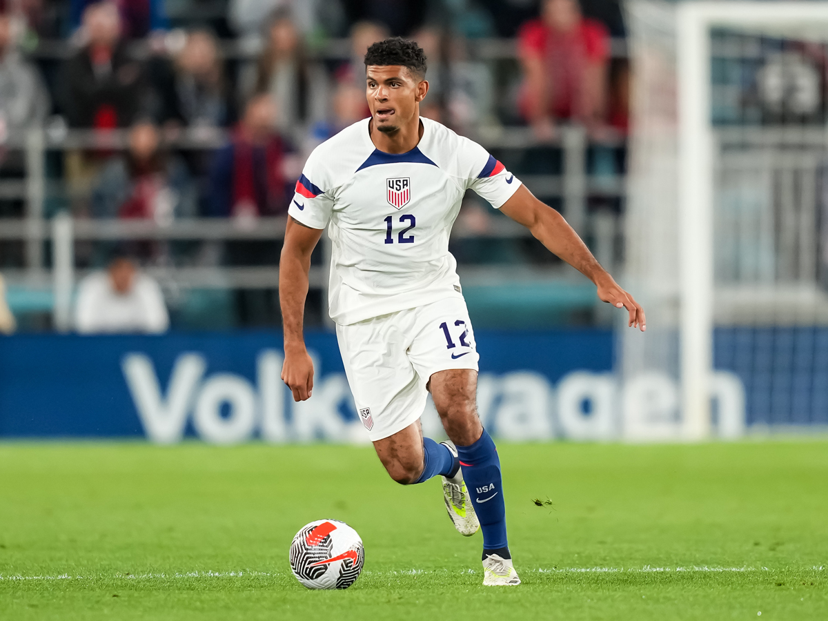 A player controls the ball during a USMNT match in his white and blue uniform.
