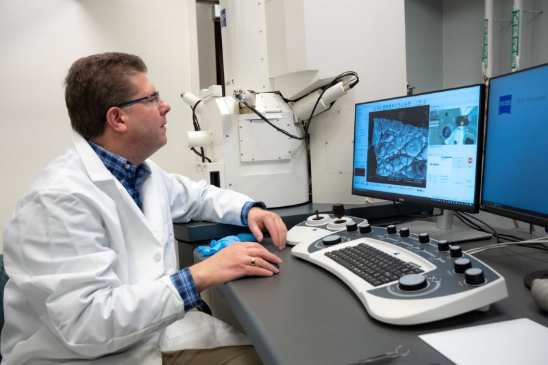 A person in a white lab coat and glasses sits at the controls of a scanning electron microscope, examining a magnified image of a textured surface displayed on a monitor in a laboratory setting.