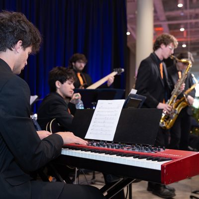 A student jazz ensemble performs at an indoor holiday event, with a keyboardist in the foreground and saxophonist and bassist behind him