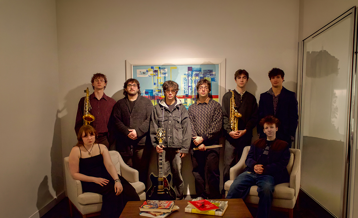 Eight members of the Orange Juice student jazz ensemble pose in a small room with armchairs and a coffee table, some holding instruments including saxophones, a guitar and drumsticks, with an abstract painting on the wall behind them.