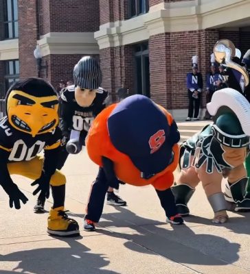 Otto the Orange lines up in a football stance alongside college mascots outside a brick building.