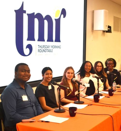 Six panelists sit at a table with microphones in front of a projected screen displaying the Thursday Morning Roundtable logo during a panel discussion.