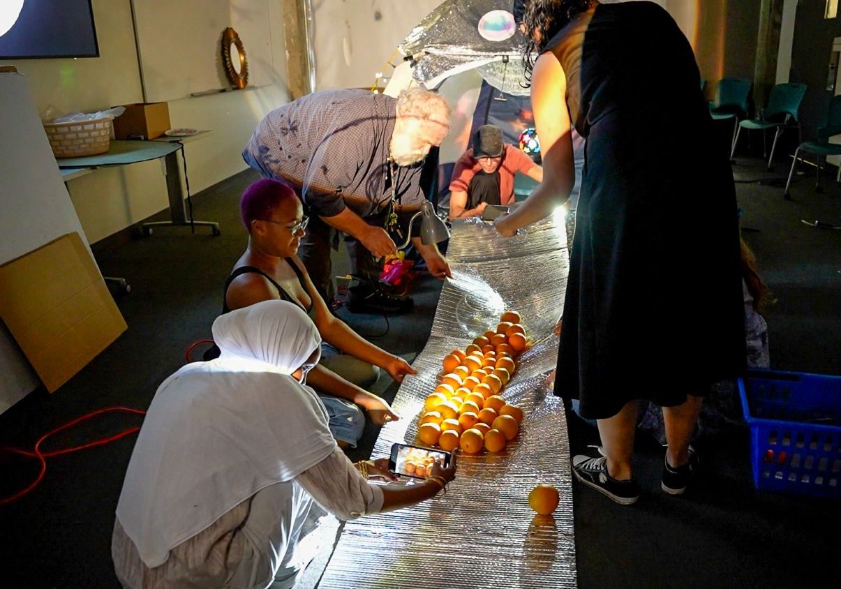 Several people gather around a reflective silver surface on the floor lined with illuminated oranges during what appears to be an art installation or film production, with one person photographing the scene with a smartphone in a dimly lit studio space.
