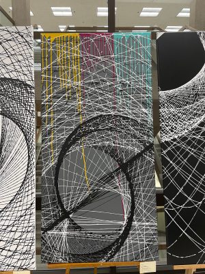 A large abstract painting on an easel featuring bold black sweeping arcs and a web of intersecting white lines over a gray background, with drips of yellow, magenta, and teal paint along the top edge.