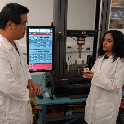 Two researchers in white lab coats discuss a mycelium sample near an Instron testing machine.