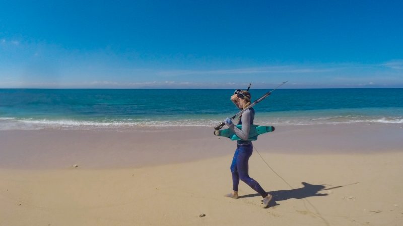 A woman in a blue wetsuit and snorkel mask walks along a sandy beach carrying spearfishing gear, including spear guns and a teal-colored float or buoy, with calm turquoise ocean water in the background.