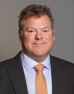 Headshot of a smiling man wearing a dark suit jacket, light blue dress shirt and orange patterned tie against a gray background