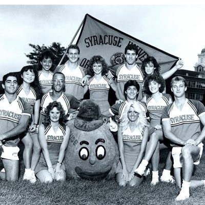 Black-and-white group photo of Syracuse University cheerleaders posing on a lawn with the Otto the Orange mascot and a Syracuse University banner in the background.