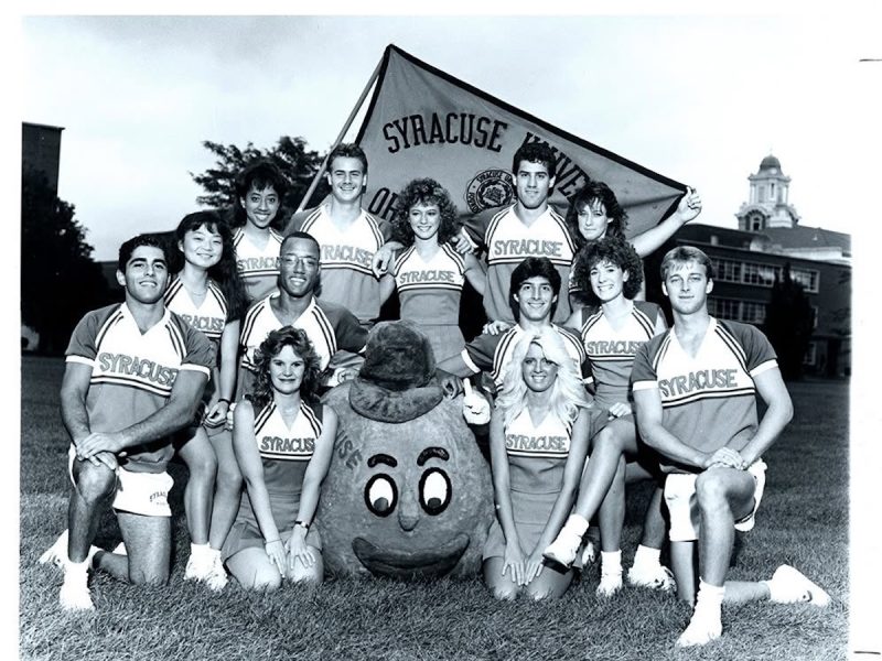 Black-and-white group photo of Syracuse University cheerleaders posing on a lawn with the Otto the Orange mascot and a Syracuse University banner in the background.