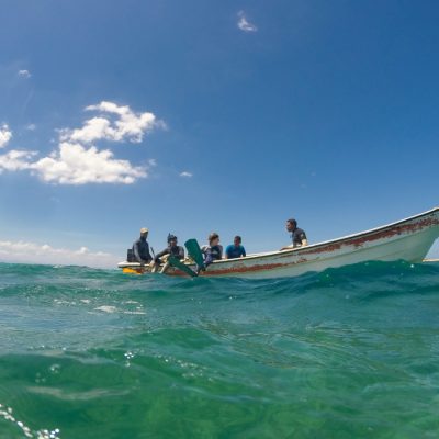 Five people sit in a weathered wooden fishing boat on open turquoise water under a bright blue sky with scattered clouds, photographed from water lev