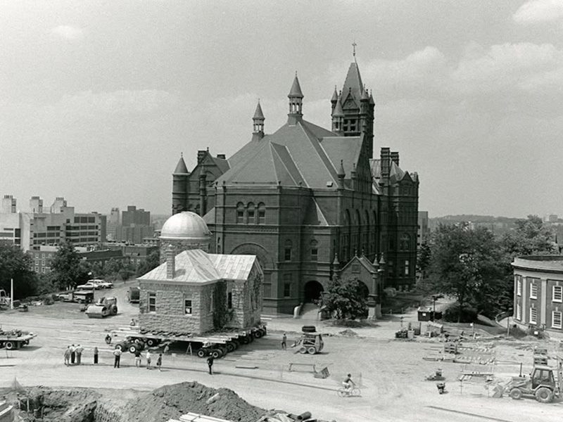 Historic observatory building being moved on a trailer across a construction site, with a large Victorian-style building in the background.
