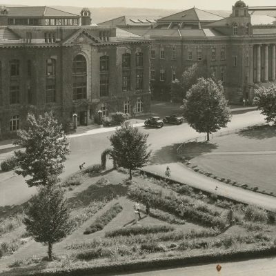 Historic black-and-white aerial view of a large ivy-covered academic building overlooking a landscaped campus quadrangle with trees, paths, and a formal garden.
