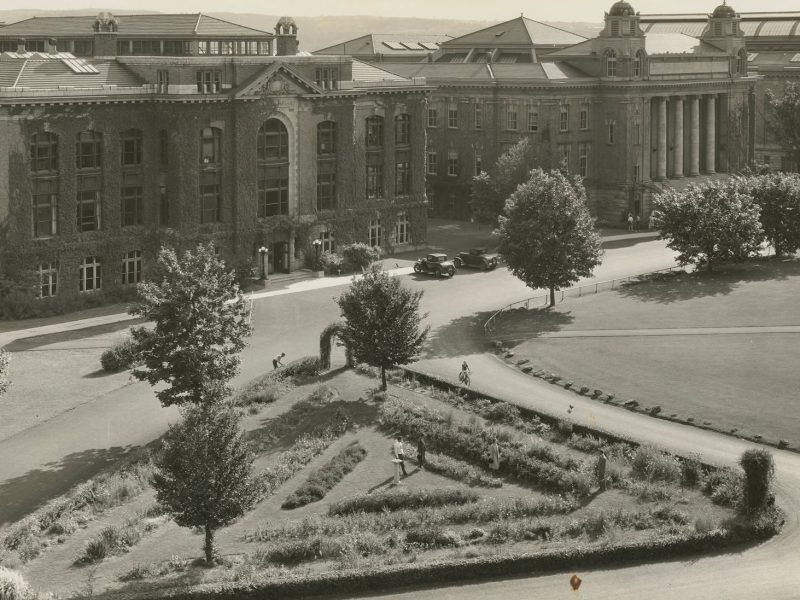Historic black-and-white aerial view of a large ivy-covered academic building overlooking a landscaped campus quadrangle with trees, paths, and a formal garden.
