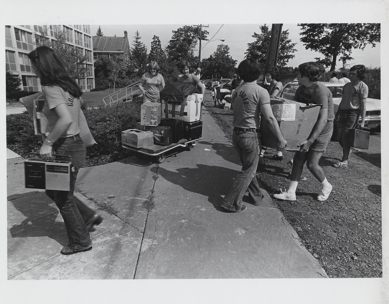 Group of students carry boxes and belongings along a sidewalk during a campus move-in day.