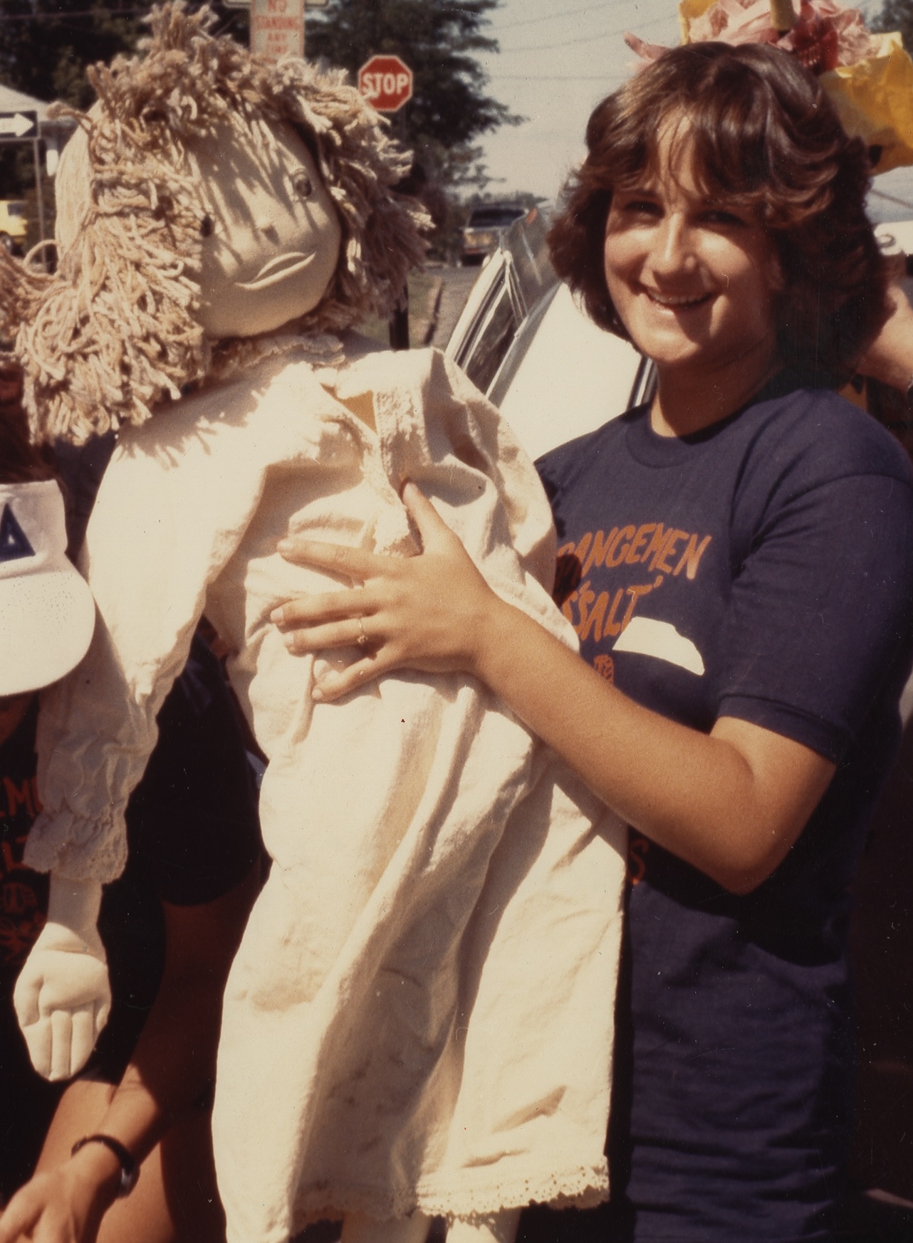 Person holding a large rag doll while standing outdoors beside parked cars.