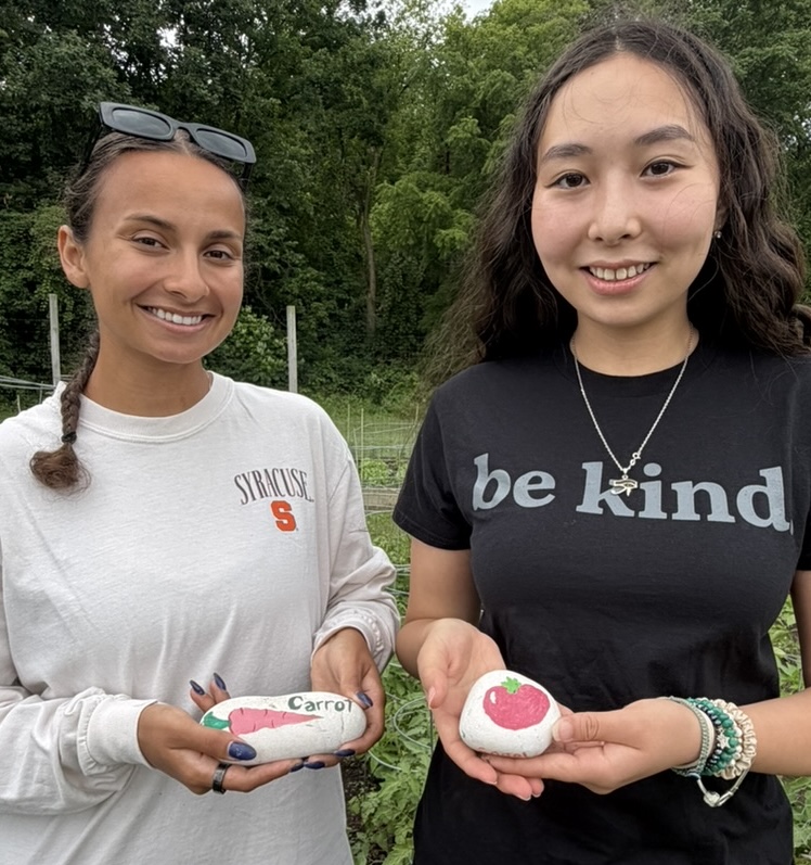 Two people in a garden holding painted rocks labeled with vegetable illustrations.