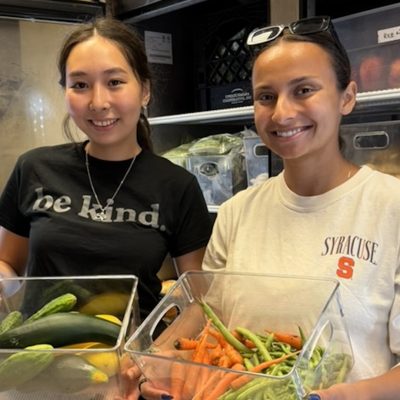 Two people stand in a walk-in cooler holding bins of freshly harvested vegetables, including cucumbers, carrots, and green beans.