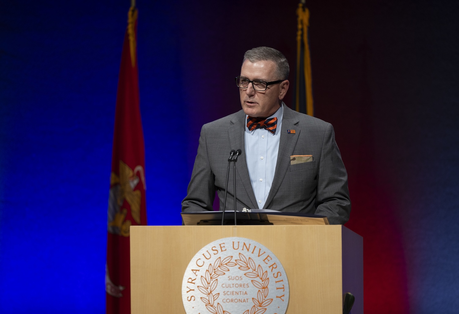 Person in a gray suit and striped bow tie speaking at a podium with a Syracuse University seal, against a blue and red stage backdrop with flags.