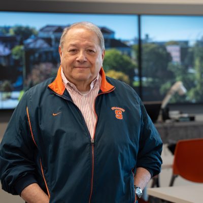 Person standing in a conference room wearing a navy Syracuse University jacket, with large display screens and orange chairs in the background.