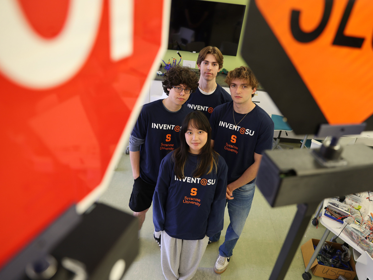 Four Invent@SU students stand near a stop sign and robotics equipment in a lab.