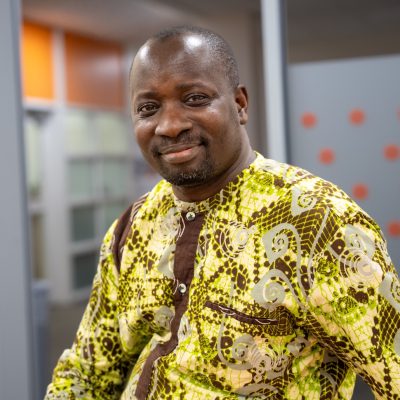 Person seated in an office, wearing a bright patterned yellow and brown shirt, looking toward the camera.