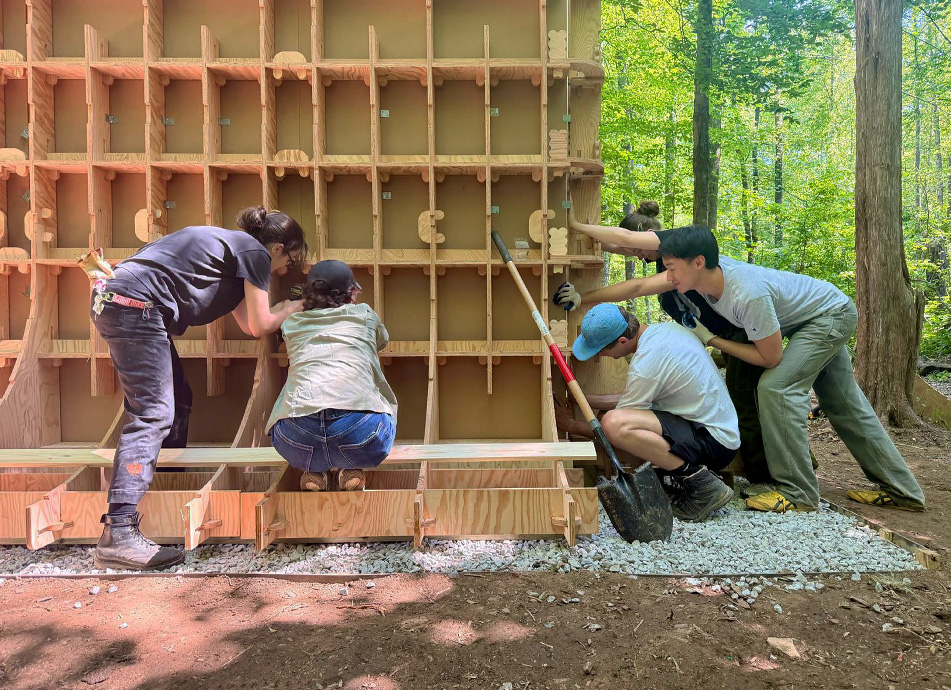 Four people work together to lift and position a large wooden lattice wall panel onto a gravel base, with a shovel propped against the structure and forested surroundings behind them.