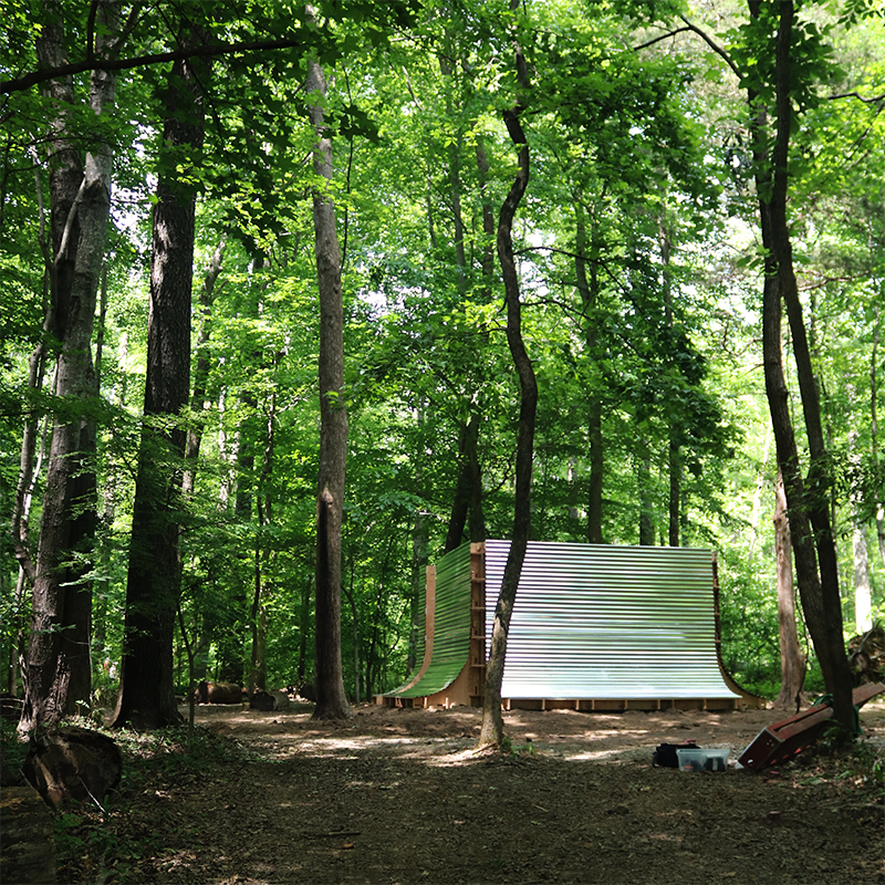 A curved wooden pavilion with an undulating slatted wall, partly clad in reflective panels, stands nestled among tall trees in a forest clearing dappled with sunlight.