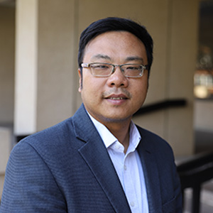 Portrait of Bing Dong, a man wearing glasses and a gray blazer over a white collared shirt, standing outdoors near a building with columns.