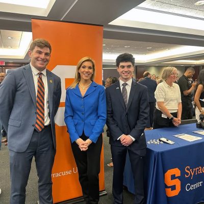 Three people pose smiling in front of a Syracuse University banner at an event.
