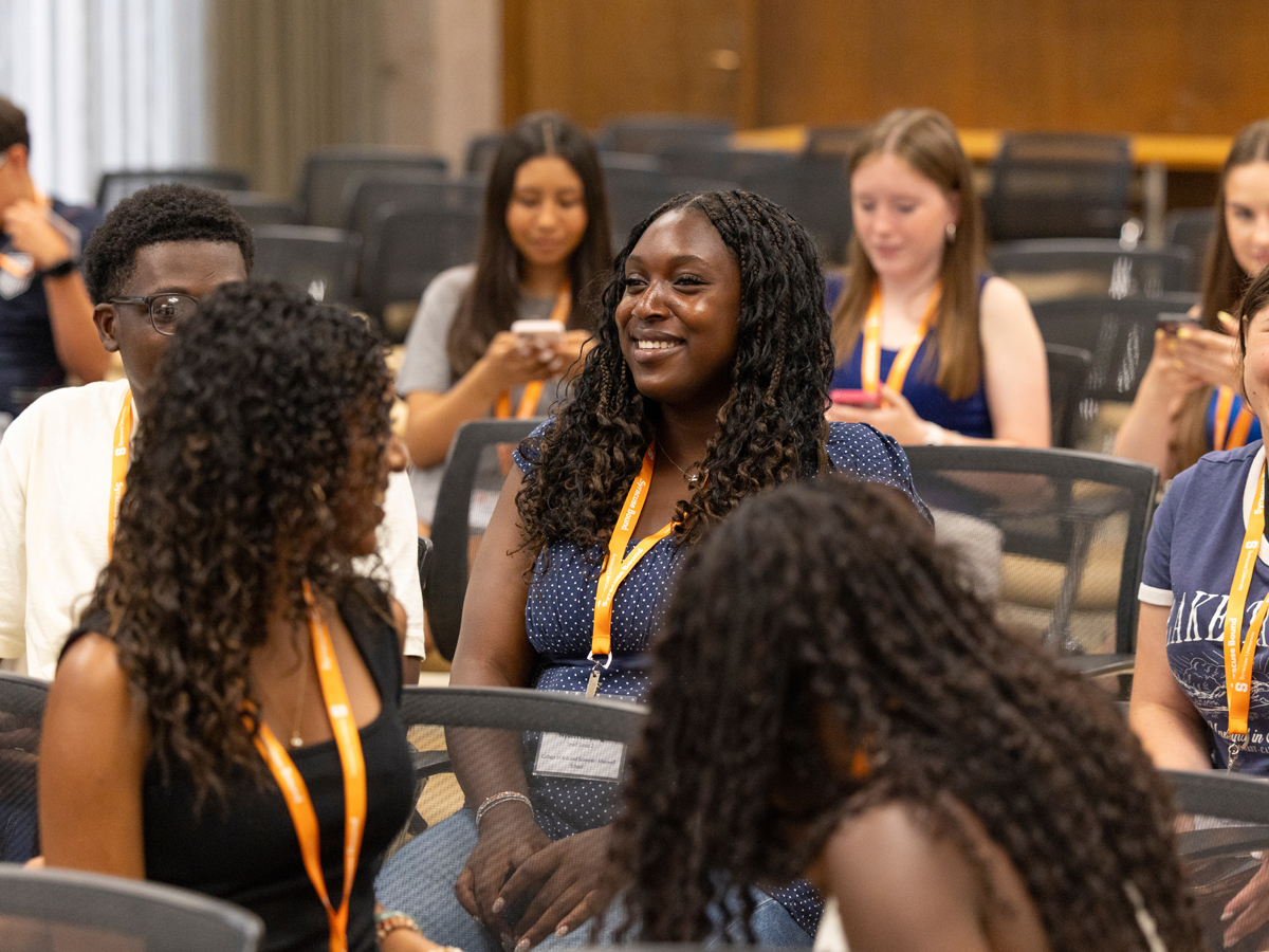 Students wearing orange lanyards sit together, smiling and talking, during an indoor orientation session.