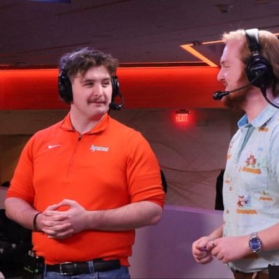 Carson Kass and Ryan Blankenhorn wear headsets, standing at Syracuse esports broadcast desk.