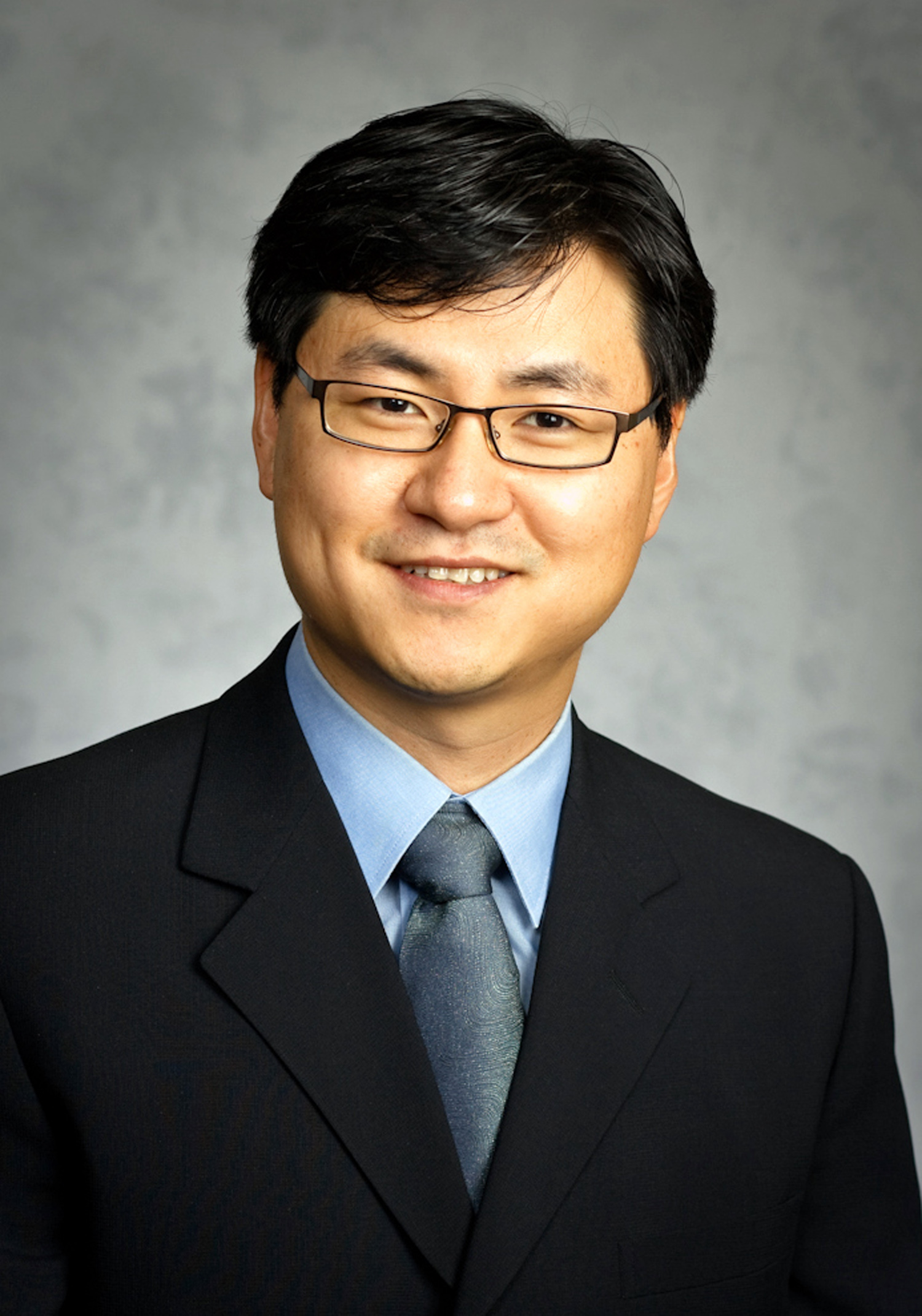 Professional headshot of a person wearing glasses, a dark suit, blue shirt, and tie against a gray studio background.