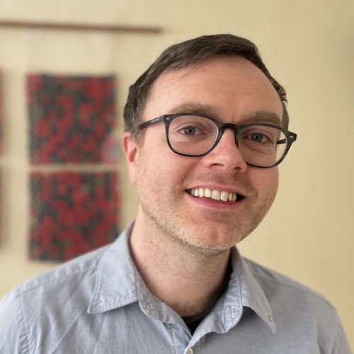 Head-and-shoulders portrait of a person wearing glasses and a light button-down shirt, with blurred artwork in the background.