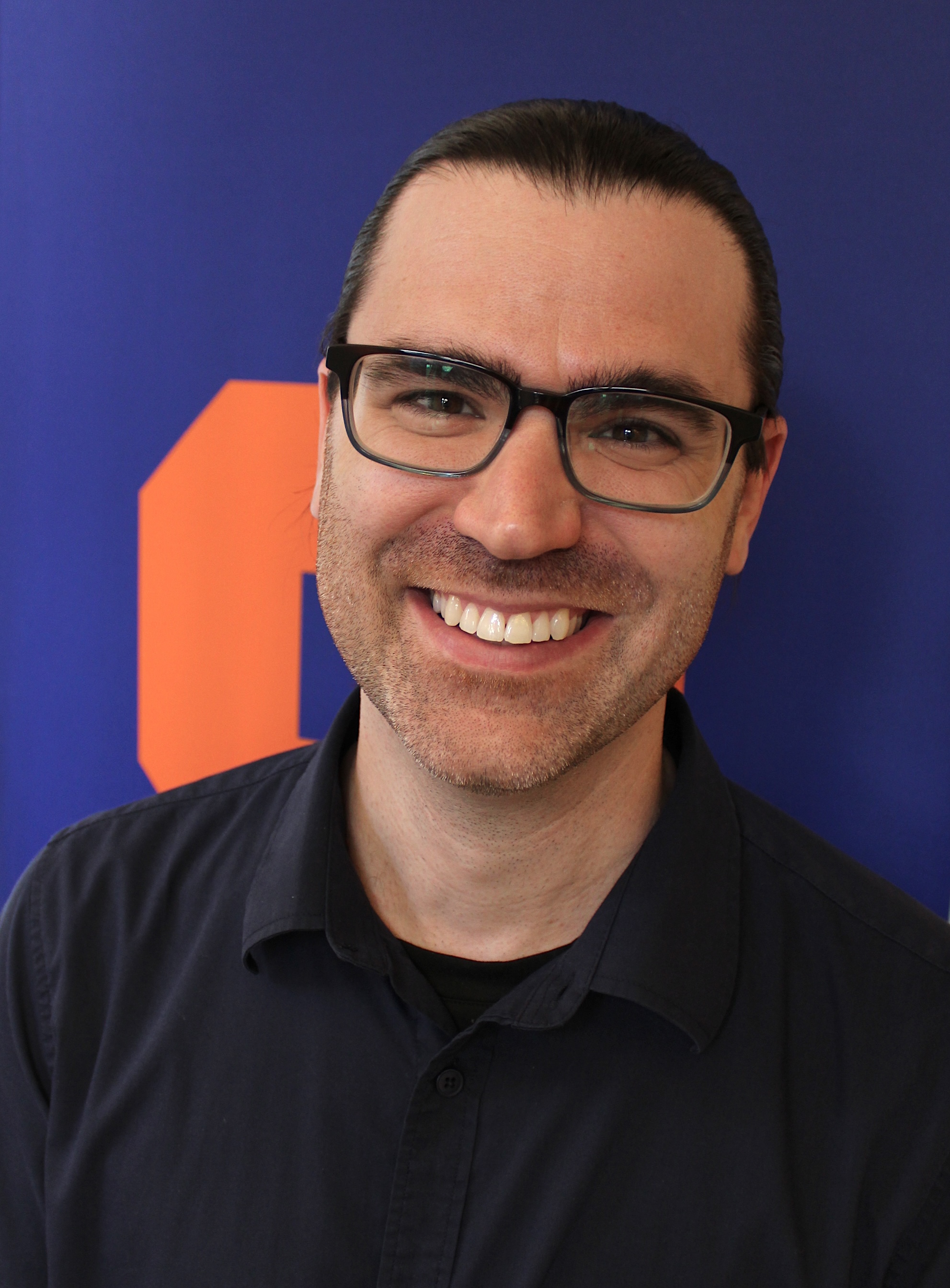 Headshot of a person wearing glasses and a dark shirt, standing in front of a blue background with an orange geometric logo.