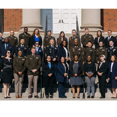 Group photo of the Defense Comptrollership Program's Class of 2026 — dozens of graduates in military uniforms and business attire standing on the steps of a brick building with white columns and doors.