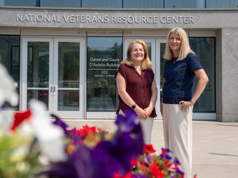 Two people stand outside Syracuse University’s National Veterans Resource Center, with colorful flowers in the foreground.