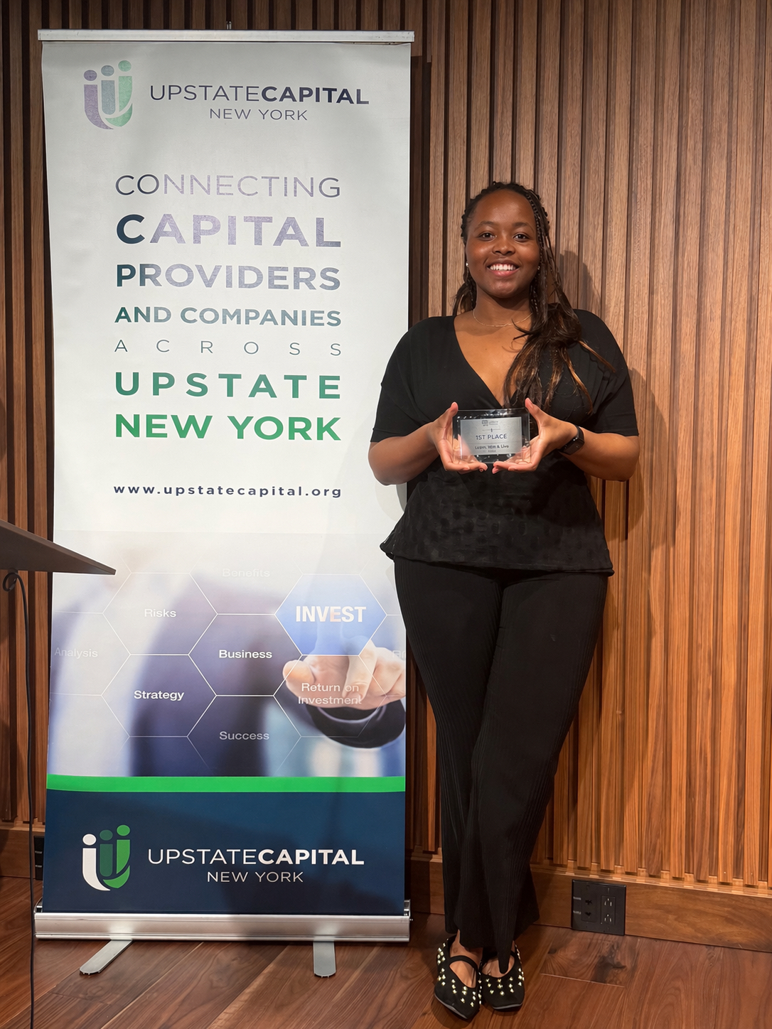Person holding a first-place award while standing beside an Upstate Capital New York event banner.