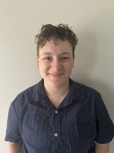 Head-and-shoulders portrait of a person with short curly hair wearing a navy patterned button-down shirt against a plain light-colored background.