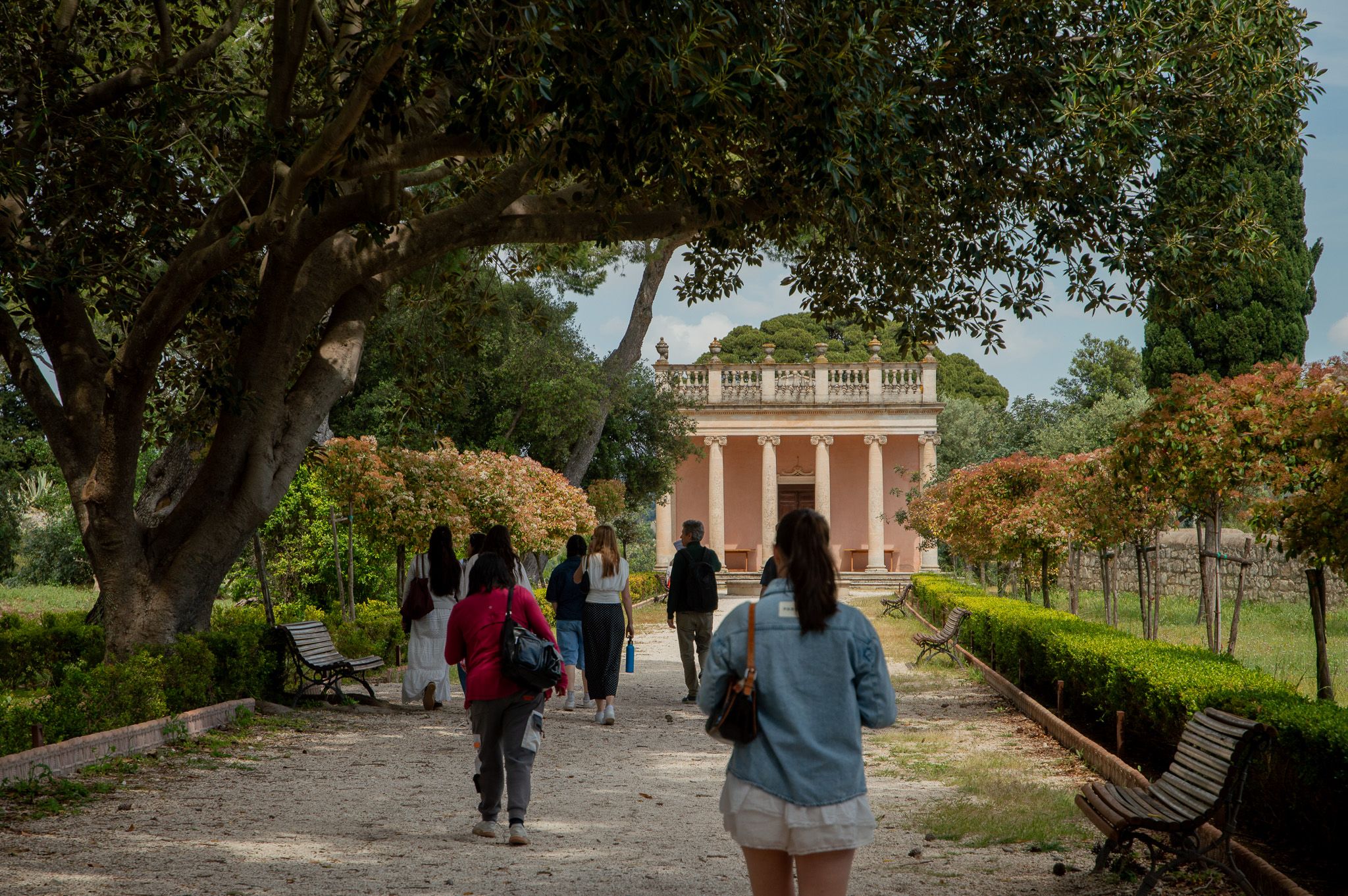 Students walk a tree-lined gravel path toward a pink colonnaded building surrounded by gardens.